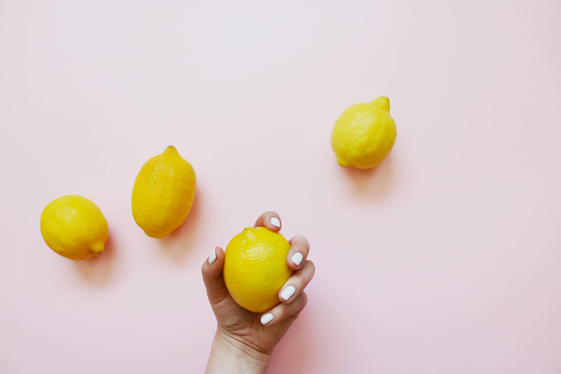 A hand with white nails holding a lemon on a soft pink background